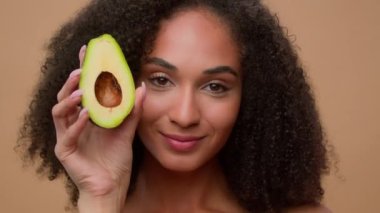 Kadın portresi stüdyosu gülümseyen Afro-Amerikan kadın elinde taze sağlıklı avokado yüz bakımı organik doğal kozmetik reklamları yapan güzel kız temiz yüz nemlendirici.