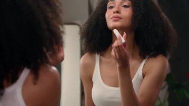 Güzel Afro-Amerikalı kadın ayna aynasına bakıyor sabah güzellik rutini banyoda etnik kız nemlendirici dudak kremi sürüyor dudak nemlendirici ruj sürüyor organik doğal kozmetik.