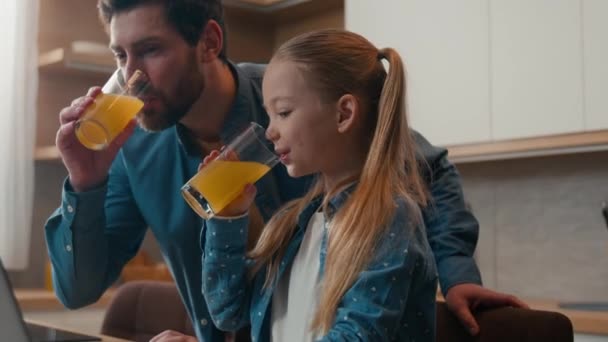 キッチン大人の父親のコーカサスハッピーファミリーと小さな娘の少女は コンピュータ技術の子供の助けを借りてラップトップの父親の教えを見て新鮮な飲み物のおいしいオレンジジュースを飲んで — ストック動画
