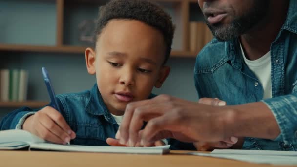 アフリカ系アメリカ人のかわいい男の子の子供のボーイで 民族的な大人の父親が宿題を書いています レッスンクラスで息子を助ける エクササイズタスク 子供の近くの耳スクーリング 小学校 — ストック動画