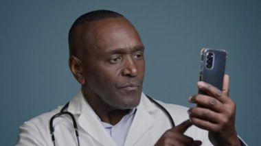 Mavi arka plan stüdyosundaki Afrikalı Amerikalı erkek doktor, telefon taraması sosyal ağları kontrol ederek akıllı telefon ekranı onay noktasını kullanarak onaylı cep telefonu e-sağlık uygulamasını onaylıyor.
