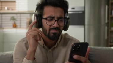 Mutlu Arap Hintli kulaklıklı adam ev mutfağında müzik dinliyor erkek milenyum dinleniyor koltukta müzik listesindeki müzik parçalarını seçiyor cep telefonundaki yüksek kaliteli ses servisinin keyfini çıkarıyor.