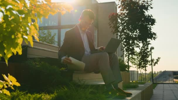 市外で働くコーカサス実業家 ラップトップの仕事 日光の大人の男教授講師は オフィスの近くで試験チェックビジネス書類の準備文書を読む — ストック動画