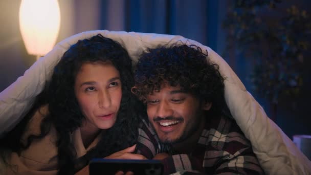 快乐的家庭男友和女友一起购物手机浏览手机软件一起笑着兴奋地观看社交媒体视频聊天一对印度男人和阿拉伯女人晚上躺在毛毯下回家 — 图库视频影像