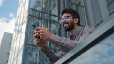 Gülümseyen Arap Hintli işadamı girişimci yönetici cep telefonu uygulamasını şehir merkezinin terasında kullanıyor alt görünüm erkek işveren sosyal medyada cep telefonuna cep telefonu mesajı yolluyor