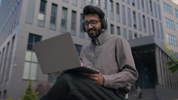アラビア人の男は オンラインで勉強するオフィスの外でビジネスプロジェクトの仕事をタイプするラップトップアウトドアで働く都市インドのビジネスマンのヘッドフォンで音楽を聞きます — ストック動画