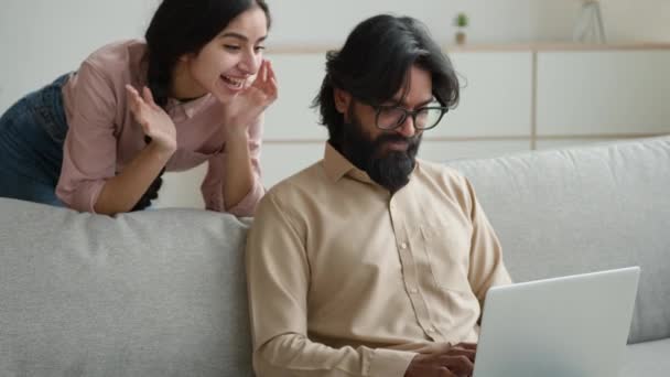 在沙发沙发上的阿拉伯裔印度人夫妇使用电脑笔记本电脑在网上商店里购物兴奋的多种族妻子丈夫快乐的兴奋的顾客女人和男人在家里的网站上搜索商品 — 图库视频影像