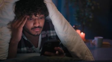 Gadget internet sosyal medya müptelası Latin Yorgun Arap adam endişeli Hintli erkek gecenin karanlığında battaniye altında yorgan kaydırmalı cep telefonuyla hayal kırıklığına uğradı