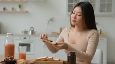 Asyalı ev hanımı Çinli Koreli Japon kız ekmeğe çikolata yapıştırıcısı sürmüş kızarmış ekmeğe tatlı reçel sürmüş çok ırklı kadın kahvaltısını evde pişirmiş süpermarketten gelen mutfak yemeklerinde sandviç yapmış.