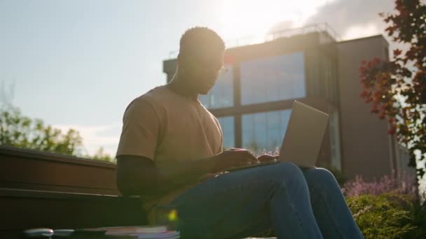 アフリカ系アメリカ人男性学生の男少年ティーンエイジャー男性を勉強する屋外大学キャンパスの近くのベンチで宿題をオンライン学習ラップトップは日光浴でコーヒーを飲むために準備します — ストック動画