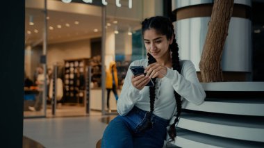 Ortadoğulu bayan iş kadını Arap kökenli kız öğrenci telefonda alışveriş yapıyor alışveriş merkezinde oturuyor mobil uygulamalar kullanıyor internetten dijital haber videoları okuyor.