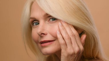 Close up mature elderly woman 50s model with pipette dripping serum vitamin C oil drops on cheek beauty face skincare hyaluronic acid moisturizing anti-aging natural liquid skin glow beige background