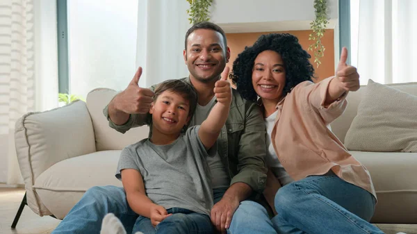Happy multiracial family African American parents embrace with Caucasian little boy son adopted kid on home floor talk laugh fun weekend smiling father mother and cute child showing thumbs up gesture