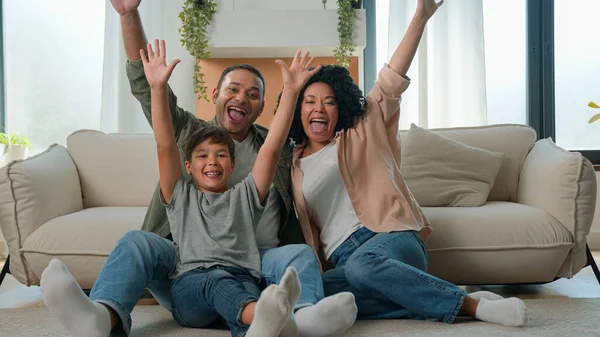 Happy multiracial family African American parents Caucasian child little boy on floor in living room own apartment mom dad kid home celebration excited father mother son raise hands together fun joy