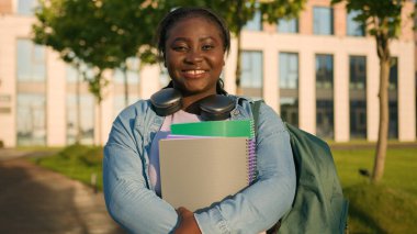 Afro-Amerikan kadın üniversitesi kampüs akademisi öğrencisinin portresi. Elinde okul çantalı kulaklıkları olan liseli kız öğrenci. Açık havada dişlek bir şekilde gülümsüyor.