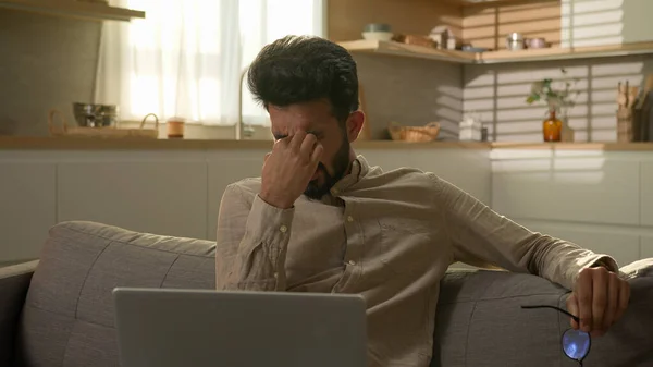 Indian Arabian man exhausted guy freelancer on couch at home kitchen working with laptop overworked suffer eyestrain eyes pain discomfort eye tension take off glasses take break bad eyesight headache