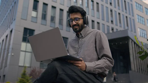 Arabian man listen music in headphones in city Indian businessman working with laptop outdoors typing business project work outside office happy student freelancer listening audio song studying online
