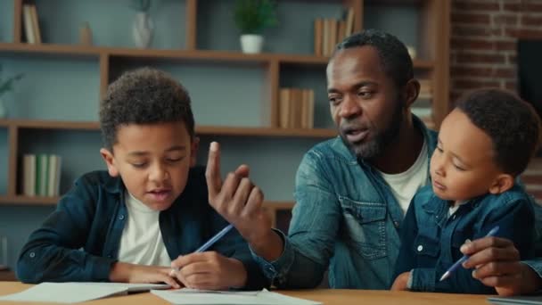 幸せな家族 アフリカ系アメリカ人の父親男性 2人の男の子の子供が家にいる子供が学校の宿題を学ぶ小学生の息子の少年を助けます 小学校の検疫学習 — ストック動画