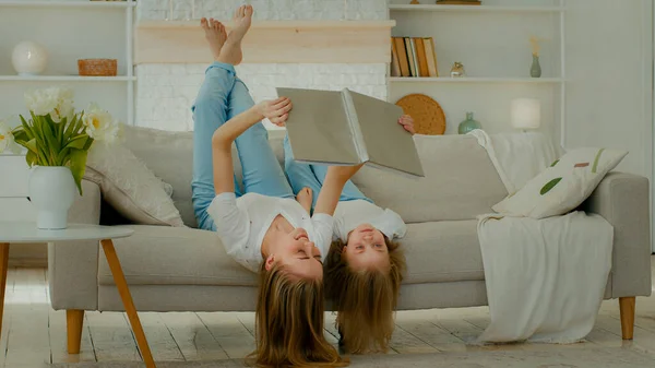 Cheerful Caucasian mother lying upside down on comfortable sofa with little child girl kid daughter reading book read together looking at pictures in textbook album family enjoy weekend at home couch