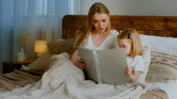 Caucasian mother read fairy tale to little daughter before sleeping at nighttime bedtime sitting in bed. Mom help preschool cute child girl kid baby reading book in at home bedroom childcare education