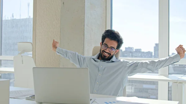 Happy office employee Arabian Indian man tired muslim businessman entrepreneur relax take break after computer laptop work relaxing relief body tension stretching arms back muscles finish job smile
