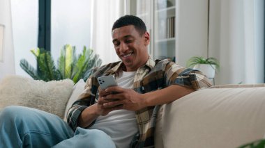 Afro-Amerikalı, mutlu, mutlu, gülümseyen bir adam, evdeki koltukta rahat bir şekilde dinleniyor. Mobil uygulama, kaydırmalı telefon uygulaması kullanıyor, sosyal ağda sohbet ediyor, internet üzerinde akıllı telefon ağı kuruyor.