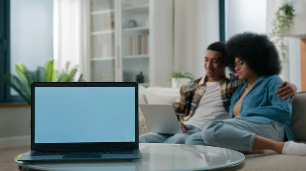 Laptop with empty copy space gray blue screen advertise ads template on foreground African American couple man woman together at home couch browsing computer shopping online insurance mortgage family