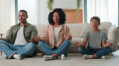Sakin, çok ırklı, barışçıl bir aile Afrikalı Amerikalı ebeveynler meditasyon öğretiyor. Gözleri kapalı çocuk yoga egzersizi yapıyor. Anne ve baba ve çocuk birlikte nilüferde rahatlayıp ev zemininde zen dengesi kuruyorlar.