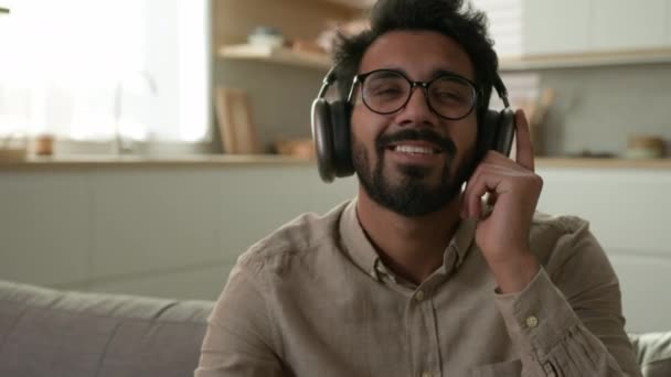 快乐的阿拉伯印度男人头戴耳机听音乐的无线耳机在家里厨房休息放松的男性房主在沙发上享受音乐歌曲在线音频服务听着声音微笑 — 图库视频影像