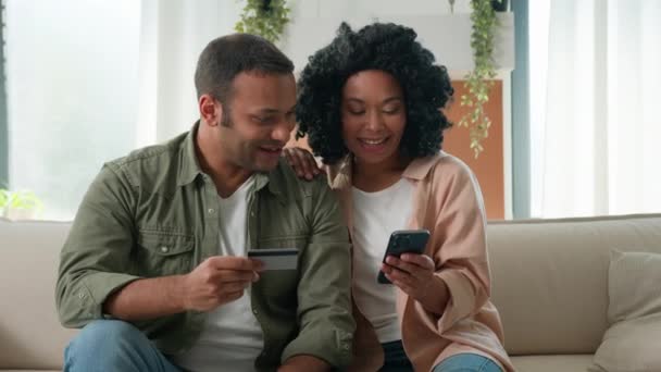 多种族的非洲裔美国人家庭在线手握手机和银行卡购物夫妇男女客户在家里预定送货付款预算快乐地看着相机微笑 — 图库视频影像