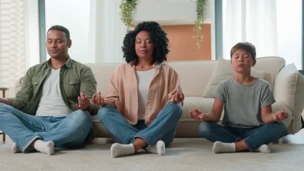 平静和平的多种族家庭非裔美国父母教静坐儿子闭目养神的孩子做瑜伽运动妈妈和孩子小男孩一起在家里放轻松荷花姿势禅定平衡 — 图库视频影像
