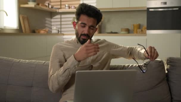 疲れたアラビアのインドの男性フリーランサーは 家庭のソファーでコンピュータのノートパソコンの仕事をオーバーワークし 眼鏡をオフにし ドライアイドライアイコン不快感の緊張を排出男性男性は痛みを感じる 悪眼の頭痛 — ストック動画