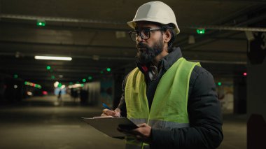 Indian contractor mechanic in helmet and glasses writing in paper examination parking lot write in clipboard. Arabian bearded man worker of manufacture work engineer in hard hat daily check inspect