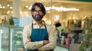 Portrait small business owner male bearded Indian Arabian man in eyeglasses standing in cafeteria restaurant posing arms crossed. Cafe waiter worker guy coffee service looking at camera crossing hands