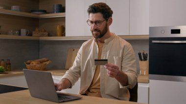 Beyaz adamın elinde kredi kartı ve internet ödemesi var. Mutfakta dizüstü bilgisayar bankacılığı uygulaması var. Web mağazası laptopunda satın almak için ödeme yapan erkek tüketici çevrimiçi alışveriş ecommerce gıda dağıtım hizmeti satın alıyor