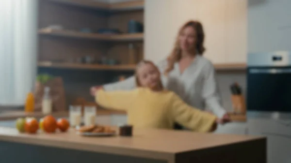 Blurred defocused view family mom and little daughter enjoy funny dance in kitchen together. Mother woman babysitter having fun with adolescent child kid girl holding hands dancing to music at home