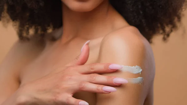 Female African American woman model beautiful girl smiling in beige studio touching perfect clean smooth healthy skin shoulder moisturizing skincare bodycare apply cream body lotion product cosmetic