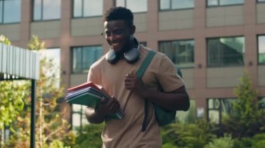 Afro-Amerikalı mutlu, gülümseyen, etnik üniversiteli erkek, sırt çantası ve kitap çantasıyla akademi üniversitesi kampüsünde eğitim dersinde açık havada yürürken gülümsüyor.
