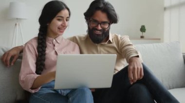 Kanepede oturan Arap Hintli çift birlikte bilgisayar alışverişi yapıyorlar. Faturaları birlikte ödüyorlar. İnternet ürünleri almayı seçiyorlar.