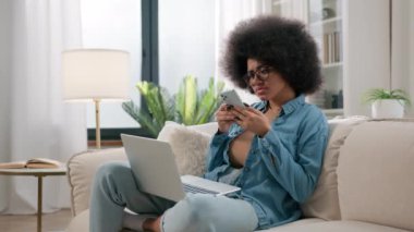 Afro-Amerikalı kadının kafası karışmış, kafası karışmış, kafası karışık, dizüstü bilgisayarlı cep telefonlu, cep telefonlu, sorunlu, sorunlu çözeltiyi seçen, evdeki akıllı telefondan yiyecek dağıtımını seçen, kafası karışık bir kız.