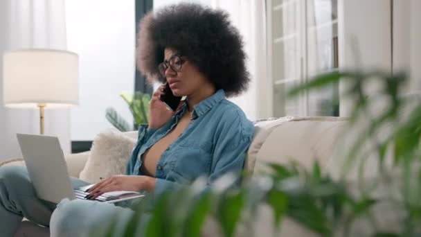 忙得不可开交的多任务非洲裔美国女商人在家里讲手机工作笔记本电脑沙发上的女客户交谈遥远的通信应答电话购物订单说话 — 图库视频影像