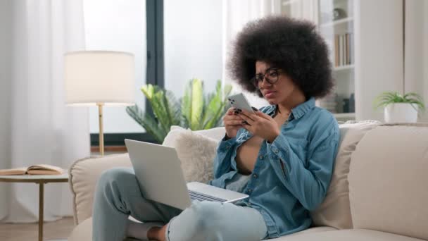 アフリカ系アメリカ人女性は混乱したペンネイティブエスニック少女を混乱させ ソファーでノートパソコンをブラウジングする携帯電話思考問題ソリューションは 自宅でスマートフォンをスクロールする遠隔作業で食品配達休憩を選択します — ストック動画