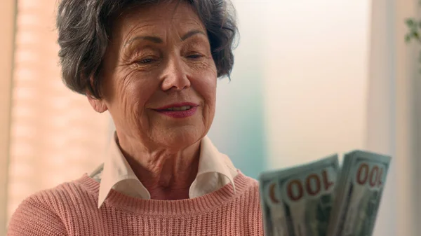 Happy smiling rich Caucasian old woman count money at home bills winning bonus grandmother counting dollars senior granny holding cash old female elderly retired lady with banknotes financial savings