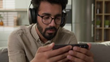 Şaşırmış Arap erkek Hintli adam video akışı izliyor online spor maç odaklı erkek kulaklık takıyor cep telefonu ekranına bakıyor ev mutfağında akıllı telefondan film izliyor