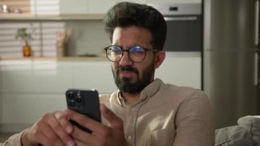 Kafası karışmış Hintli Arap bir adam. Kanepedeki erkek ev sahibi. Akıllı telefon aleti kullanıyor. Sosyal medyayı okuyor.