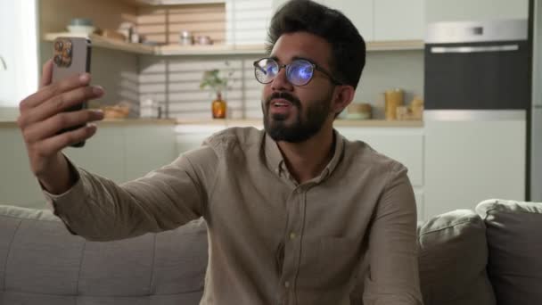 笑顔で陽気なインドのアラビア人男性エスニック男フリーランサー幸せなブロガーホームオーナーは キッチンでソファーでオンラインブログの会話をチャットするスマートフォンとのビデオ通話携帯電話の距離ミーティング — ストック動画