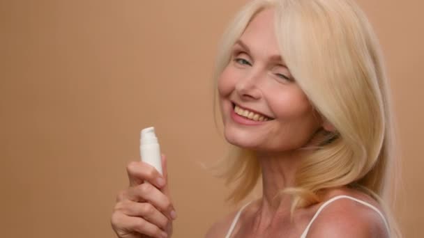 在米色演播室里笑着年长的女人白种人成熟的老太太拿着一瓶面霜润肤霜防老化提神化妆品天然美容防皱胶原蛋白豪华护肤广告 — 图库视频影像