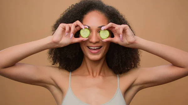 Beauty routine happy young girl beautiful attractive African American woman with curly hair holding fresh cucumber slices circles eyes cover posing beige studio background organic cosmetics skin care