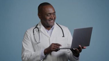 Afrikalı Amerikalı erkek doktor terapist. Mavi arka plan stüdyosunda beyaz önlüklü erkek cerrah. Bilgisayarda hastalara danışmanlık yapıyor.