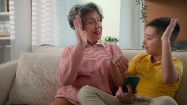 ハッピー面白いおばあちゃんと祖母は 家庭のソファーで一緒に携帯電話を再生すると スマートフォンのチームワークの成功でオンラインゲームで勝利を祝う少年との高い5人のハンドグランドマンを与えます — ストック動画